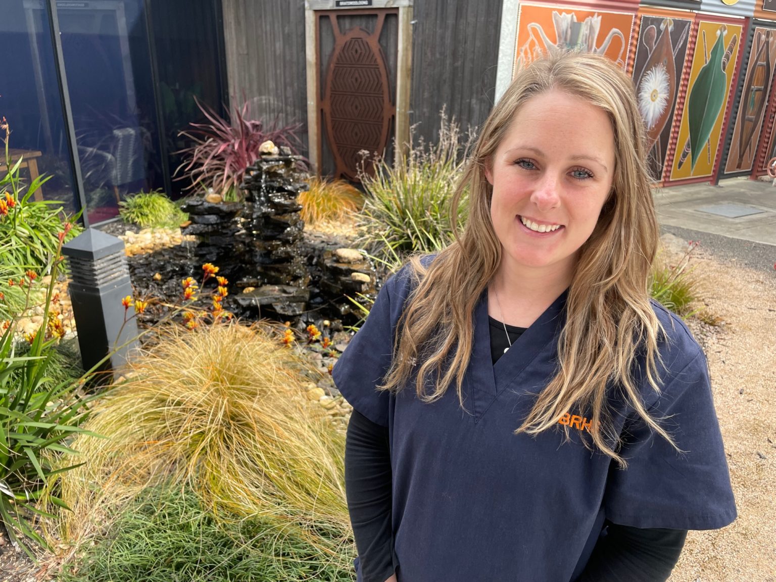 Ashley Leach is wearing a dark blue BRHS uniform and is standing in front of a garden with a water feature outside the BRHS Oncology and Dialysis Unit.