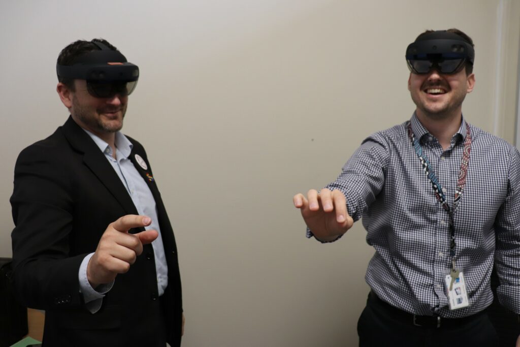 Darin Roy, left (wearing a blue BRHS jacket), teaches Orbost Regional Health's Mitch Schwenke to use HoloLens. Both men are standing in an office and wearing bulky, dark virutal reality goggles across their eyes and are pointing at a virtual object they can see through the goggles.