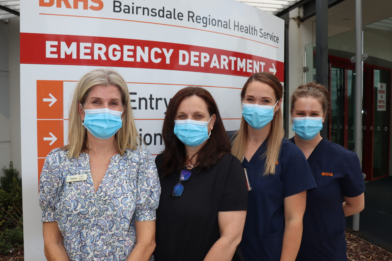 Four women, each wearing surgical masks, stand side by side in front of a BRHS sign directing visitors to the Emergency Department.