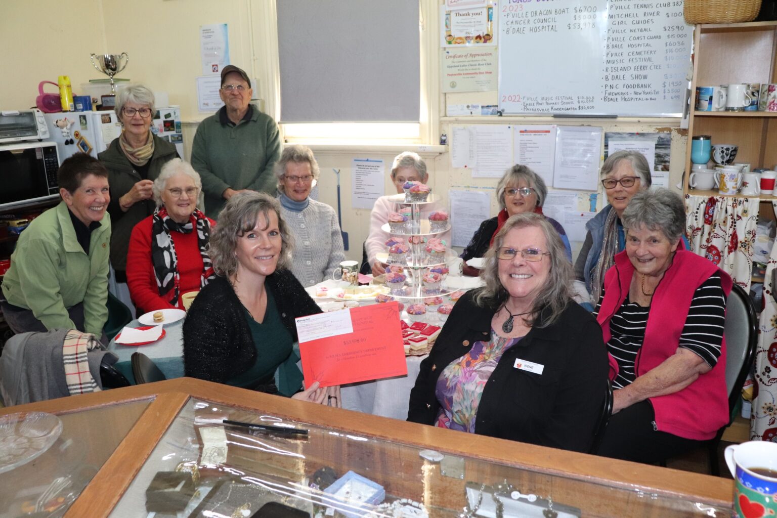 Eleven people gather around a table that has cakes and slices for an afternoon tea. One of the people is holding a cheque to donate to Bairnsdale Regional Health Service.