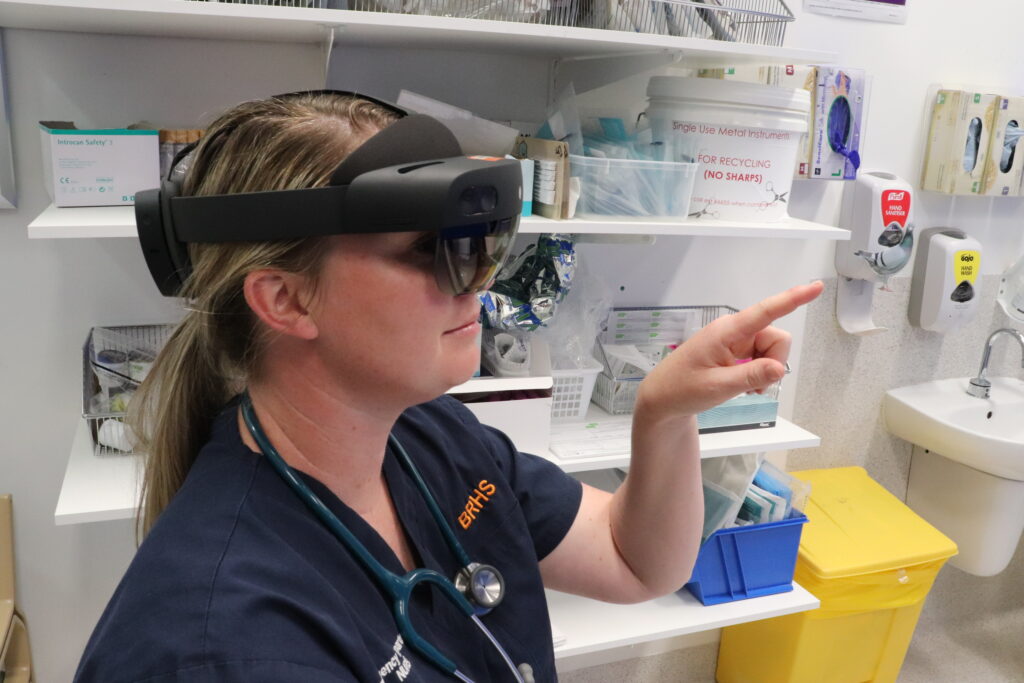 A nurse in a dark blue BRHS uniform is wearing a set of HoloLens augmented reality goggles and is pointing her left index finger forward.