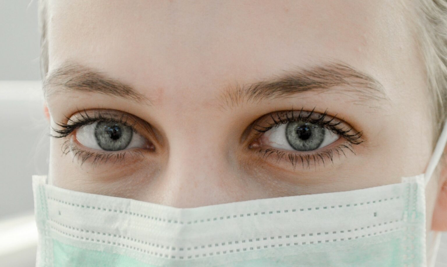 A woman wears a surgical mask and cap. Only herblue eyes are visible.